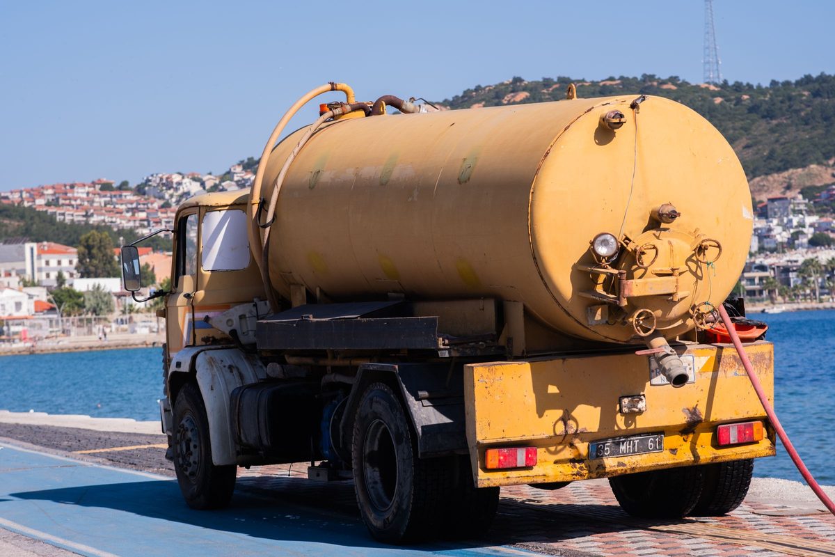 Yellow tank truck with a suction hose, parked on a waterfront road